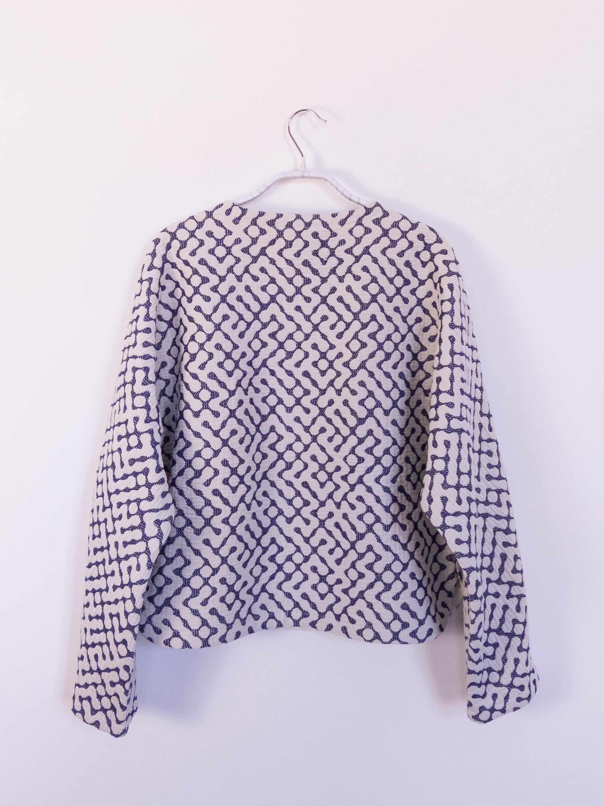 Rückansicht der handgefertigten „The Orb Awakens Jacket“ aus Baumwolle, blau mit weißem kreisförmigem Muster, auf einem Haken hängend.