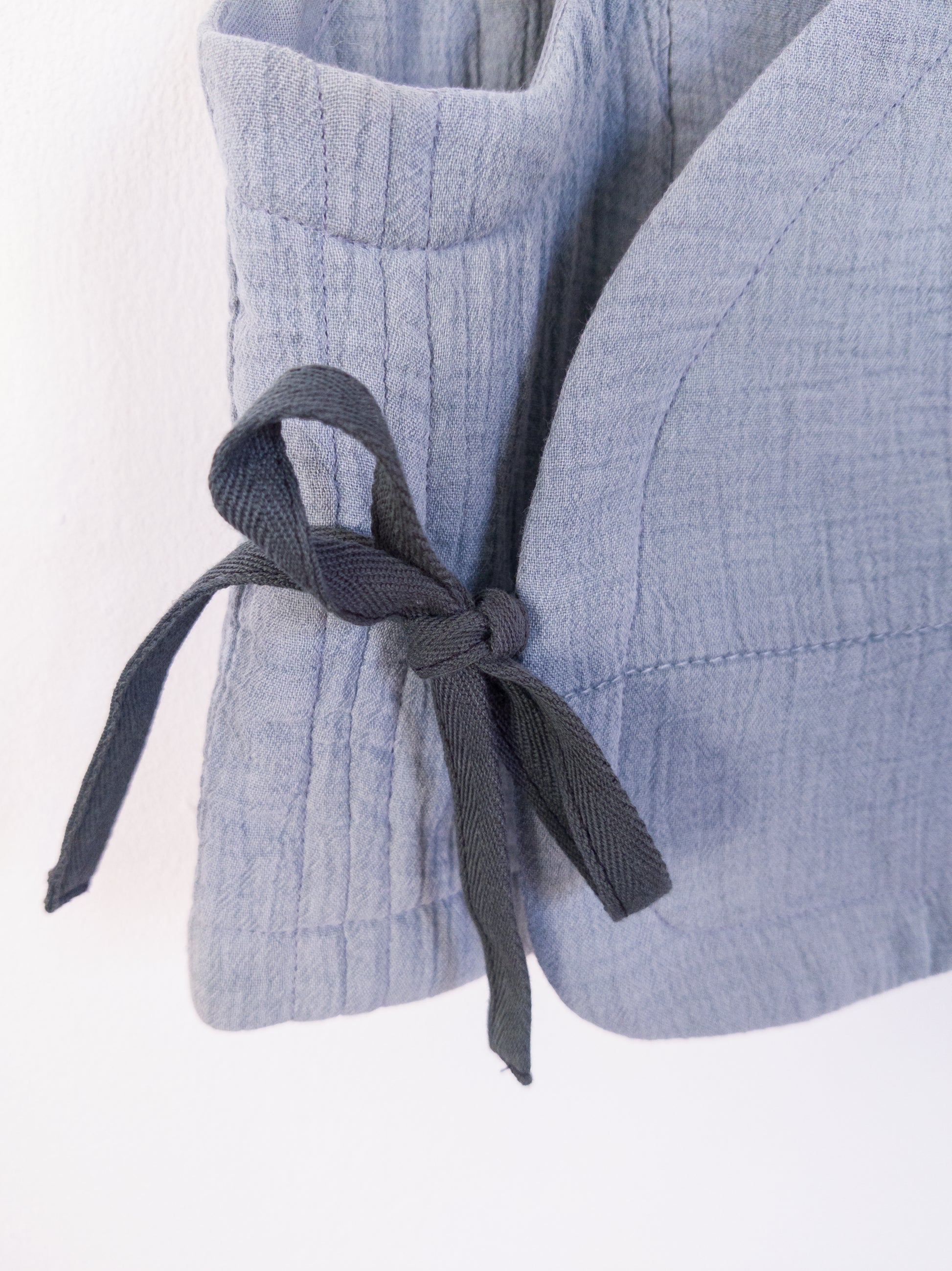 Detailansicht der „Soft Spot Vest“ zeigt die weiche, gesteppte Musselinstruktur in Blau, aus Baumwolle mit Polyesterfüllung, gebunden mit grüner Schleife.