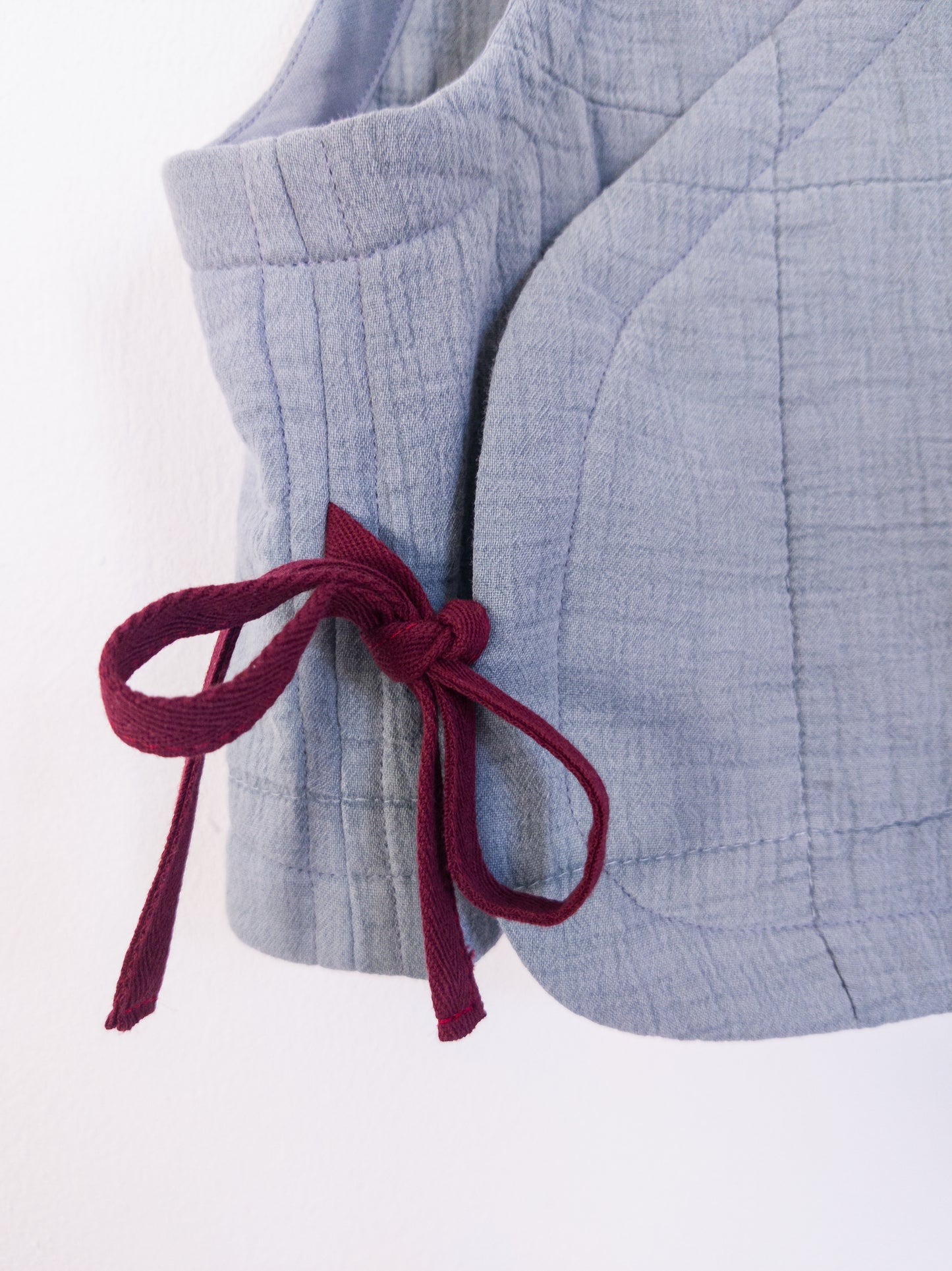 Detailansicht der „Soft Spot Vest“ zeigt die weiche, gesteppte Musselinstruktur, gebunden mit roter Schleife.