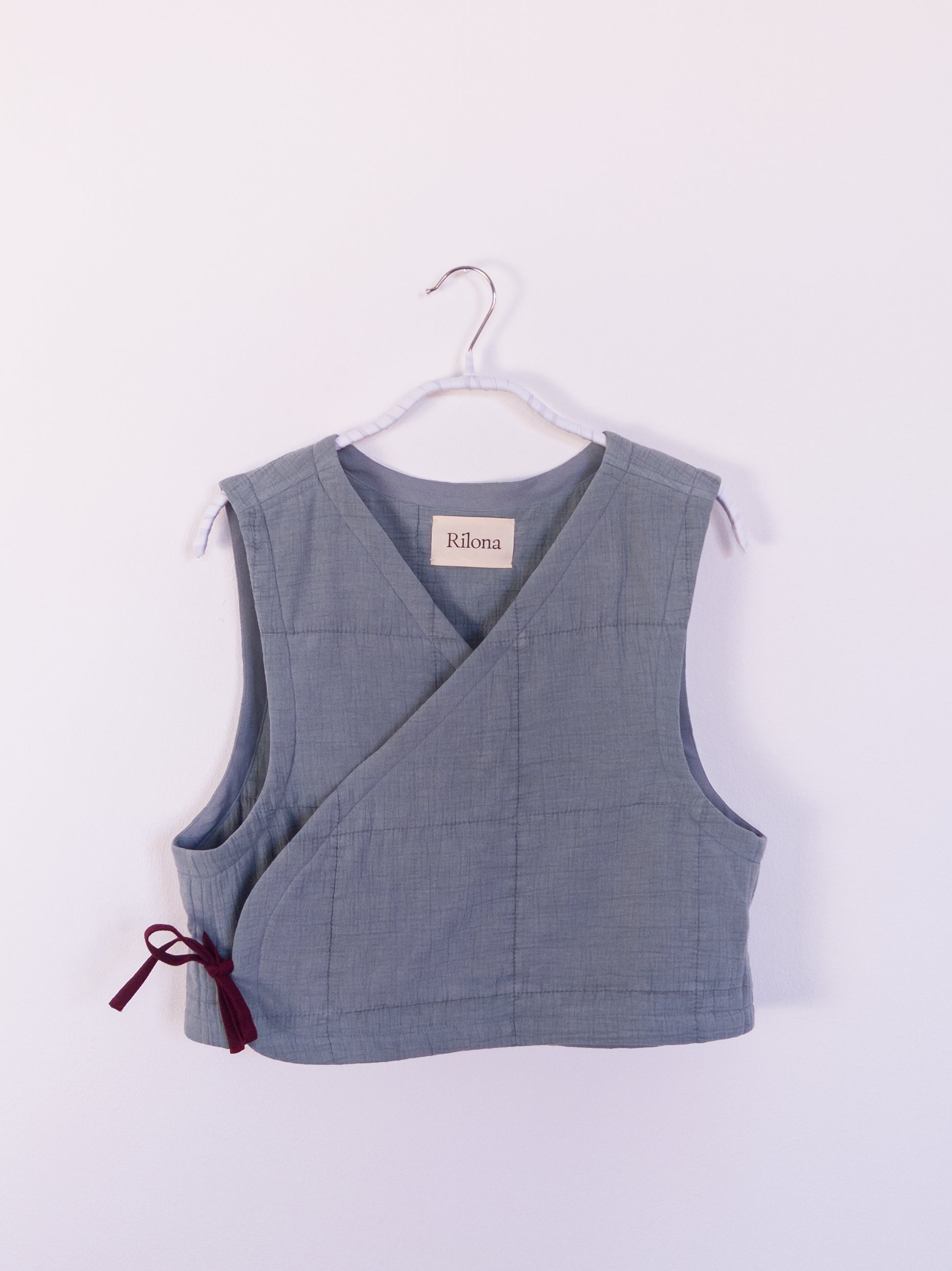 Vorderansicht der handgefertigten „Soft Spot Vest“ aus blauer Musselindecke mit quadratischer Steppung, gebunden mit roter Schleife.